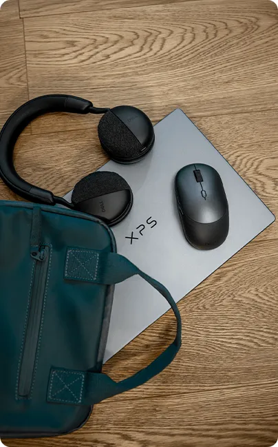 Zamknięty laptop XPS 13 leżący na drewnianej podłodze, obok słuchawki nauszne, myszka i torba